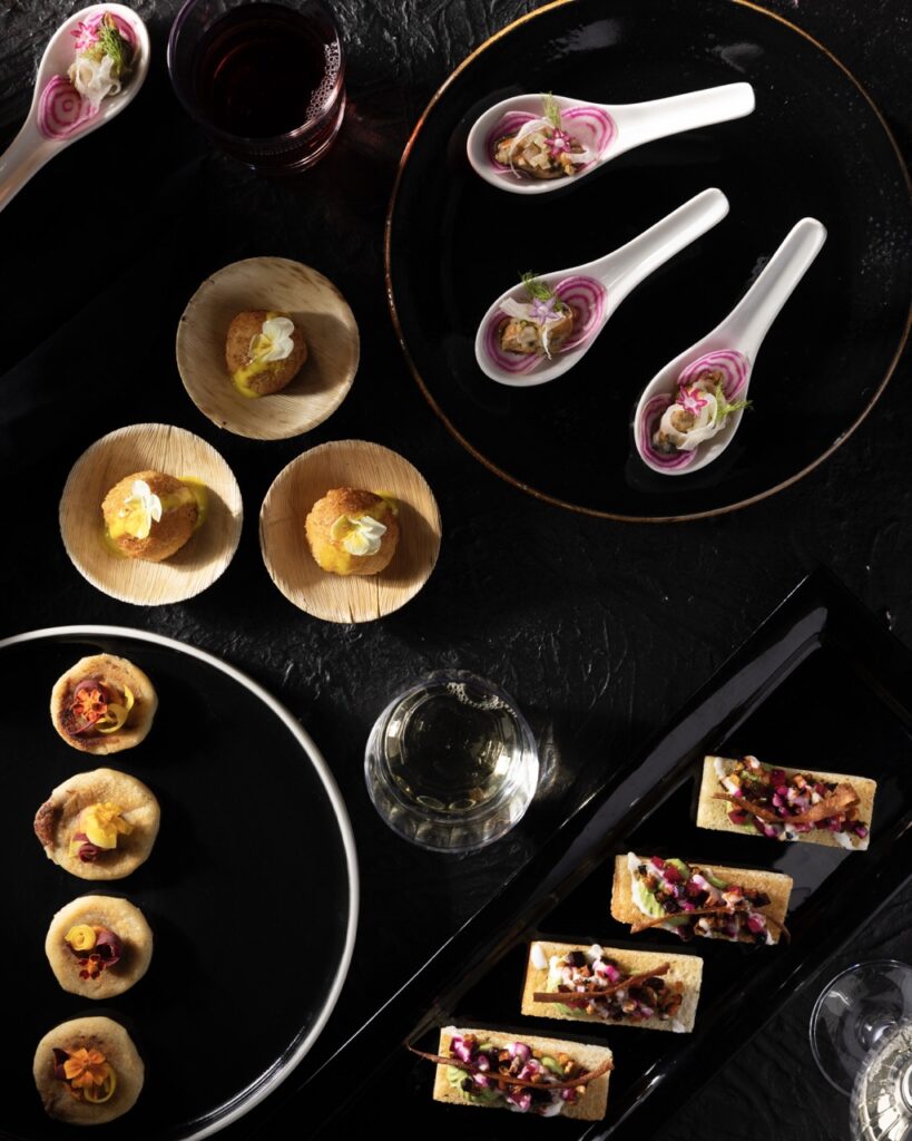 A collage of four white spoons with fresh veggies in them, three wood bowls with crab cakes and white and yellow flowers, four round appetizer bites on a black plate, and four cracker appetizer bites on a rectangular black tray.