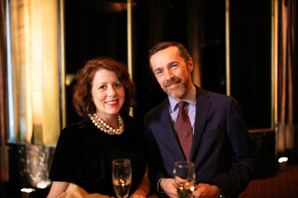 A man in a dark blue jacket holds a glass of wine next to a woman in a black dress with white pearl necklaces.
