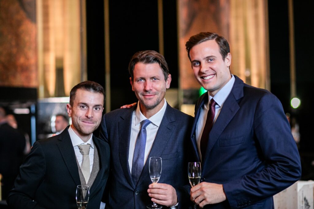 Three men, two in blue suits and one in black stand with smiles and wine glasses in hand.