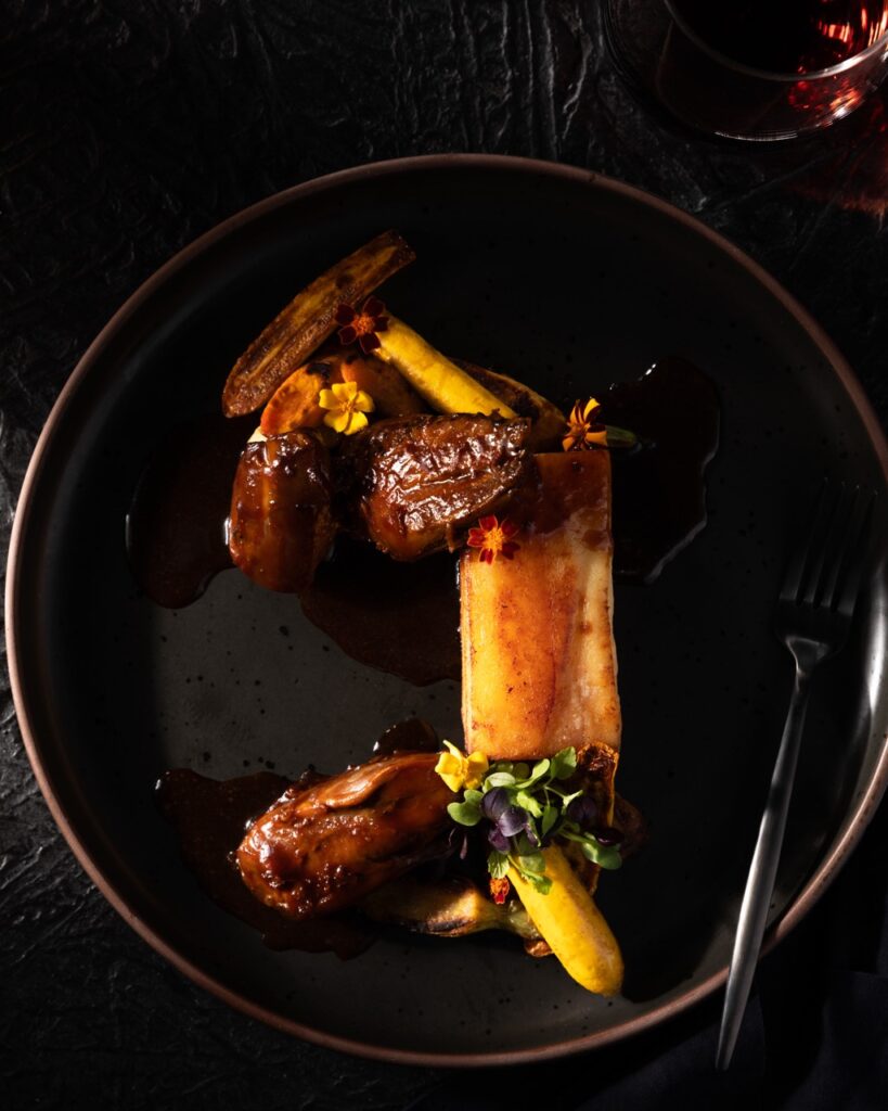 On a black plate sits roasted quash blossoms with tiny edible flowers in red, yellow, and green placed on top delicately.