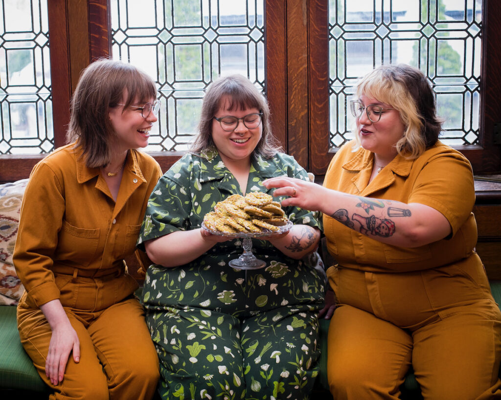 Three bakers sit in front of a window with a plate of pistachio wreath cookies in front of them