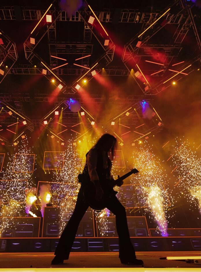 A person playing guitar on a stage with big lights and sparks from the ceiling.
