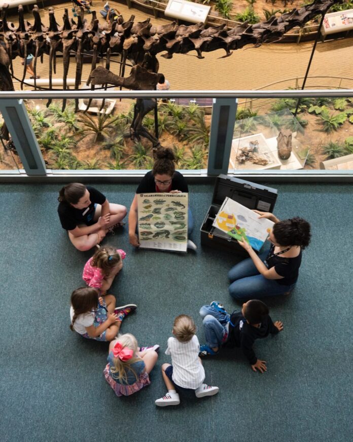 A group of kids sit on a floor in a circle with a woman reading a book to them in front of a dinosaur model.