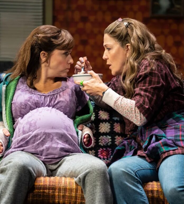 A woman holds a bowl of soup out to another pregnant woman sitting on a couch in the play Kimberly Akimbo coming to Pittsburgh.