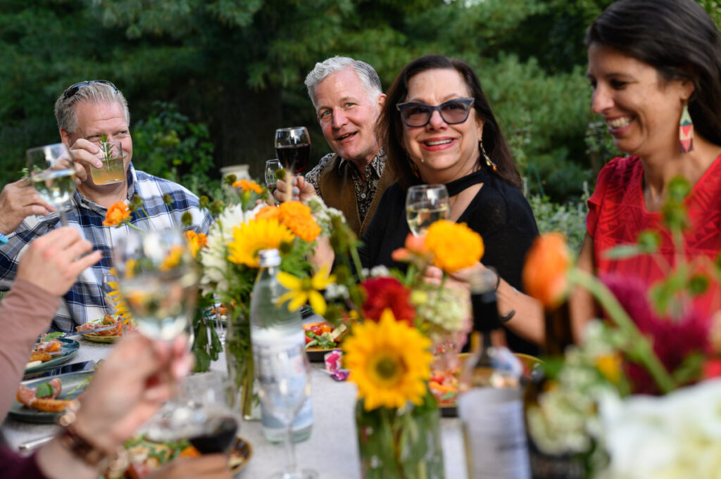 A group of people gathered outdoors in a lush, green backyard setting, enjoying a farm-fresh meal and drinks together.