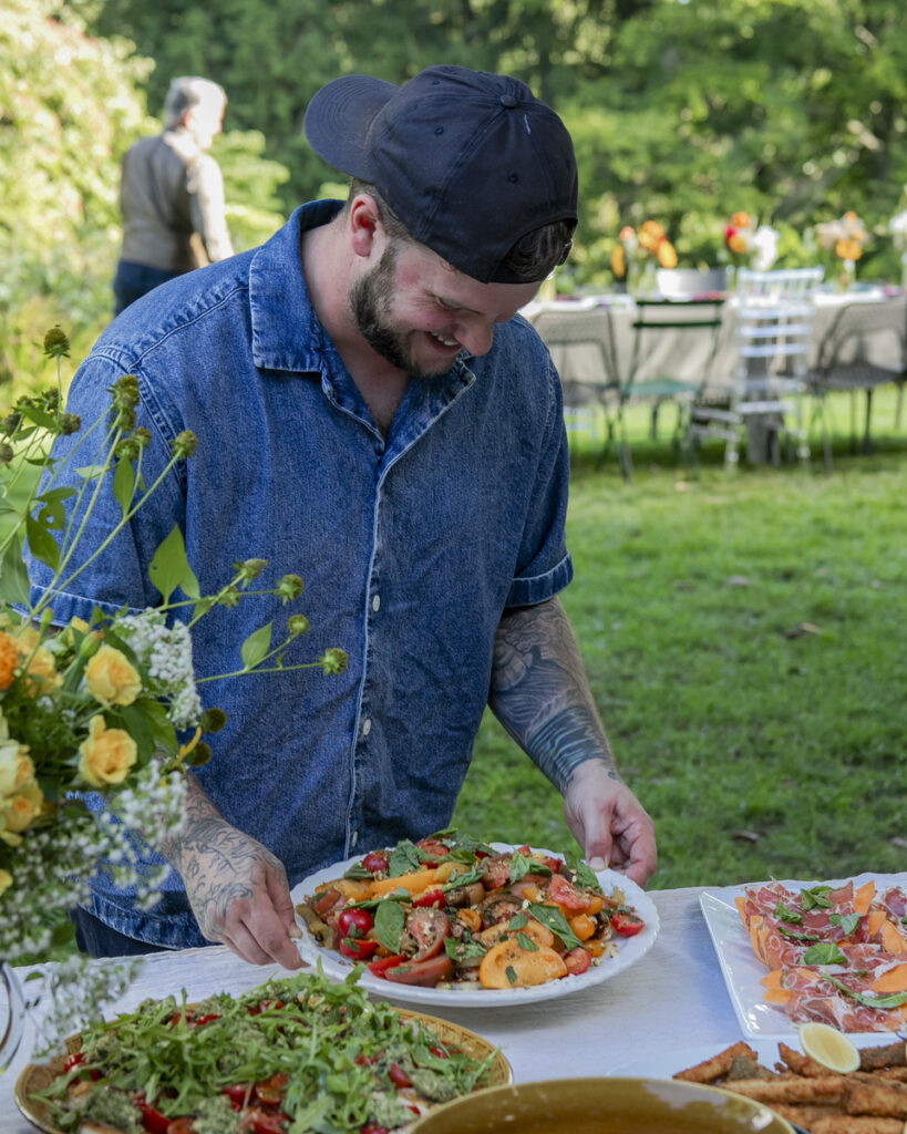 A man, dressed in a denim shirt, carefully arranging and plating a colorful, fresh salad on a table set in an outdoor garden setting.