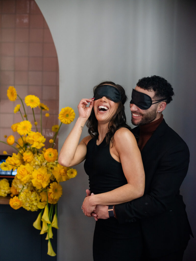 A smiling couple, both wearing dark blindfolds, standing amidst a vibrant floral arrangement, embodying the concept of exploring senses beyond sight.