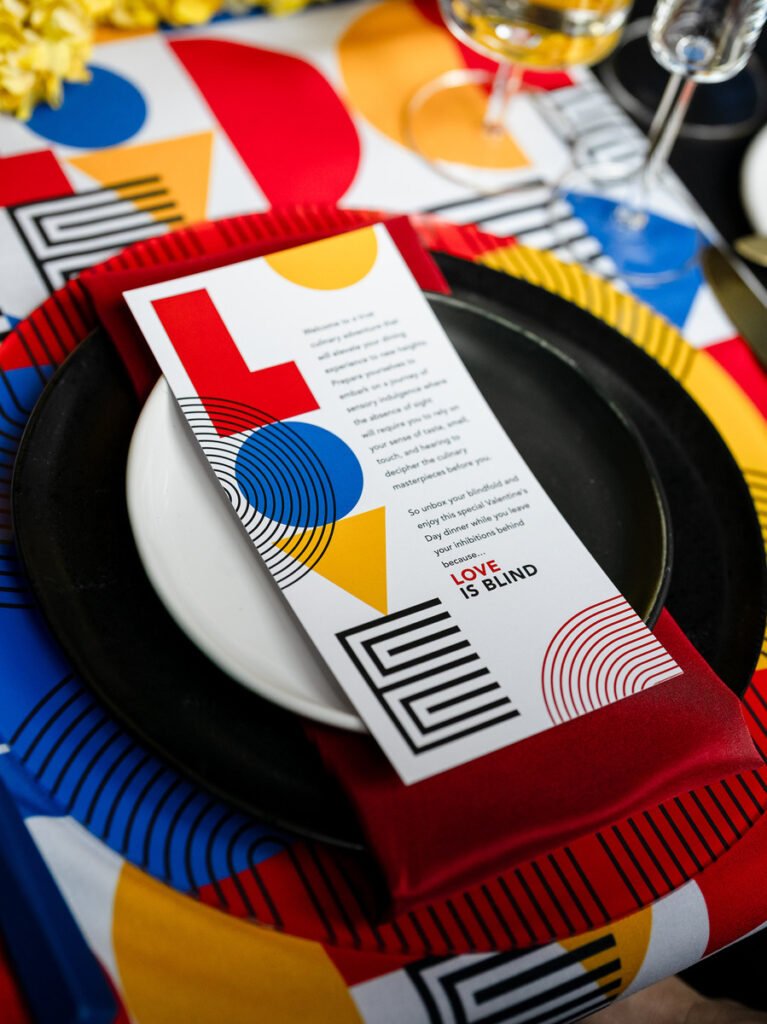 A table setting with a bold, geometric-inspired stationery item featuring the text 'Love is Blind', surrounded by a vibrant, colorful arrangement of flowers, tableware, and glassware.