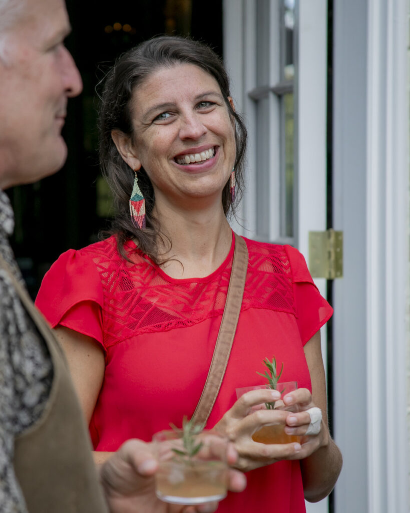 A woman in a bright red, patterned top smiling and conversing with another person while holding a drink in her hand.