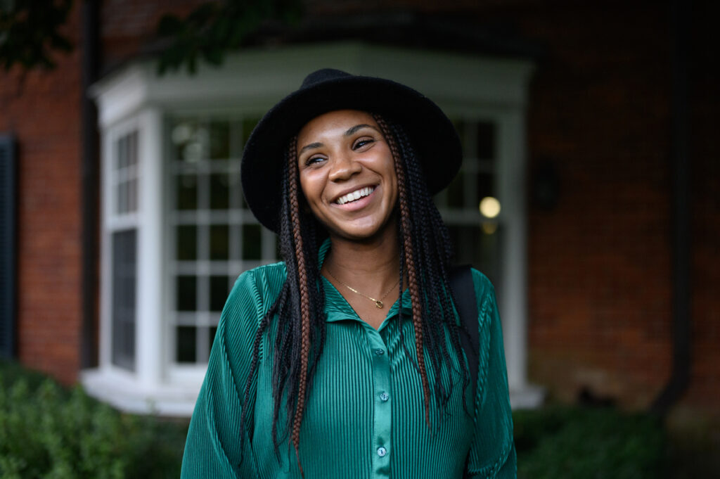 A young woman with an infectious, joyful smile. She is standing in front of a building with a warm, inviting expression, suggesting a comfortable and relaxed atmosphere.