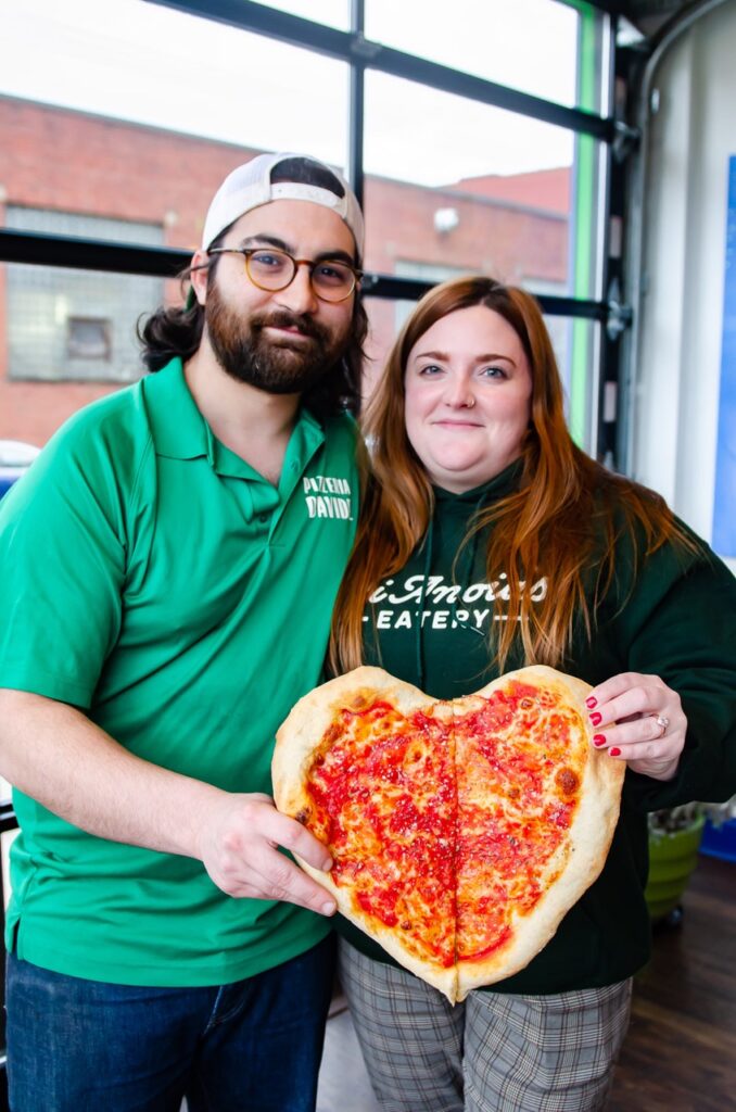 A man and woman hold a heart shaped pizza.