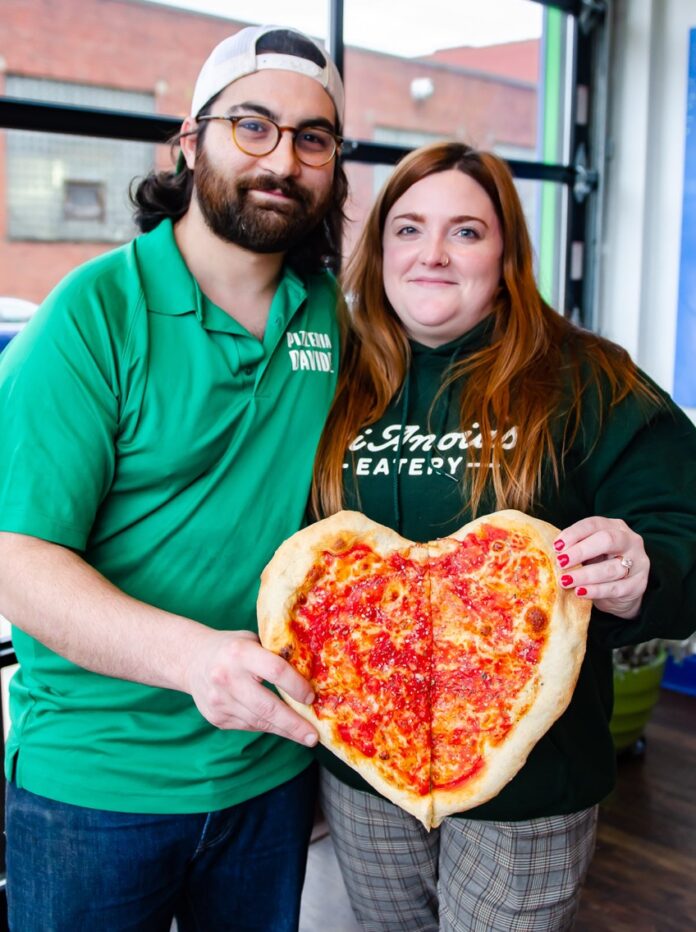 A man and woman hold a heart shaped pizza.