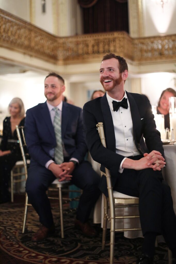 Two men in black suits sit in chairs, looking back at the front of the room.