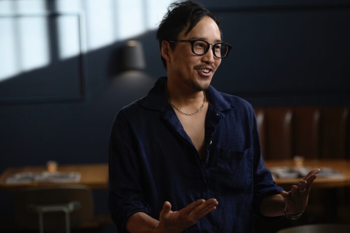 A man in a blue button up shirt and glasses talks using his hands to a crowd.