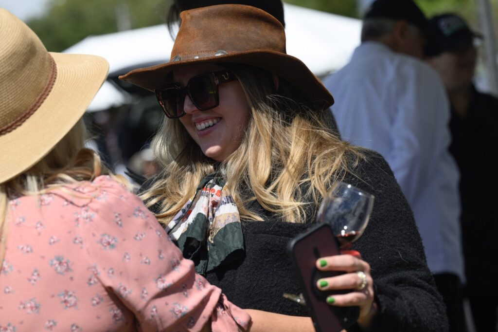 A woman in a brown leather hat and sunglasses smiles and greets another woman in a pink dress.