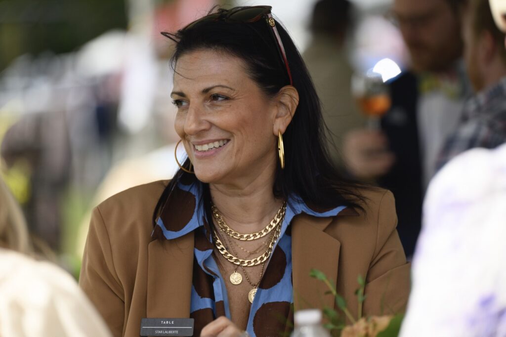 A woman in a blue shirt and brown jacket smiles off frame with sunglasses on the top of her head.