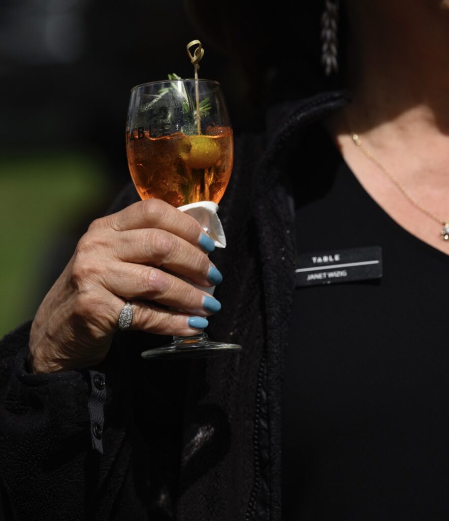 A person in a black shirt and blue painted nails holds a wine glass with a sprig of thyme and lemon inside.