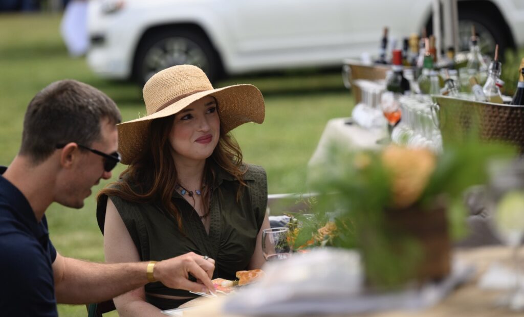 A woman in a brown sun hat and green dress sits at a table outside next to a man in a blue shirt with sunglasses.