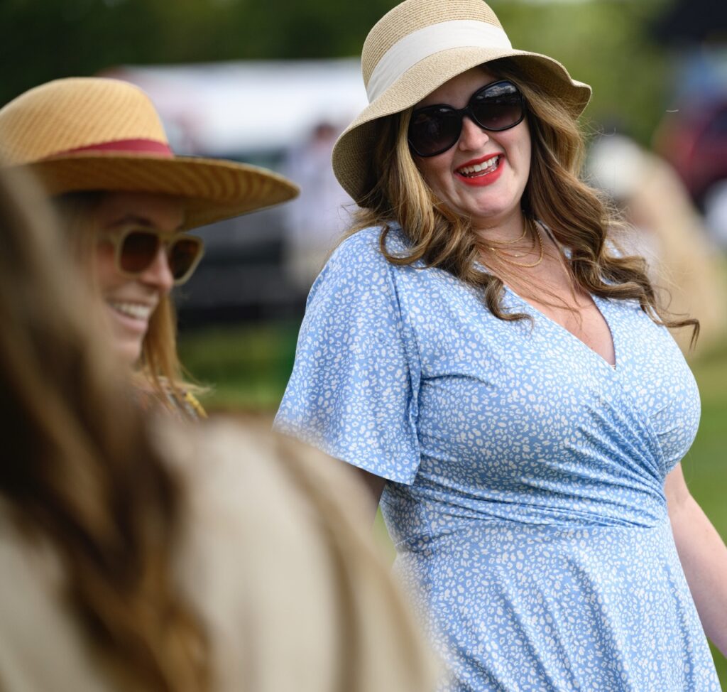 A woman in a blue sundress, brown hat, and sunglasses smiles next to a woman in a brown hat and sunglasses.