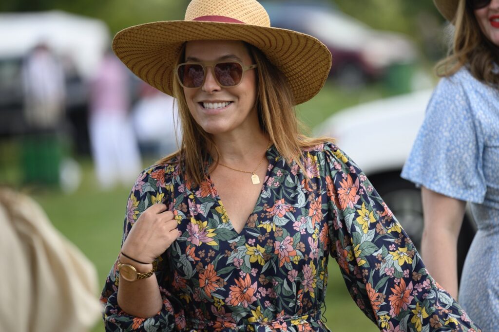 A woman in a brown hat with a red band around it smiles in brown sunglasses and a floral, colorful dress.