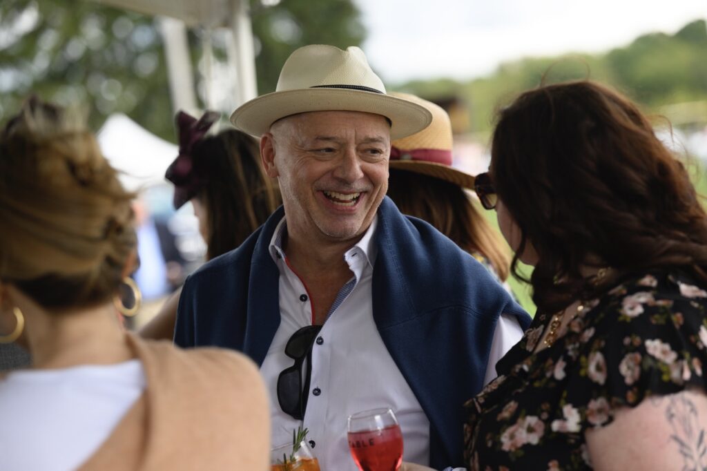 A man in a beige hat and blue vest over his button up shirt smiles at a woman with brown hair and sunglasses.