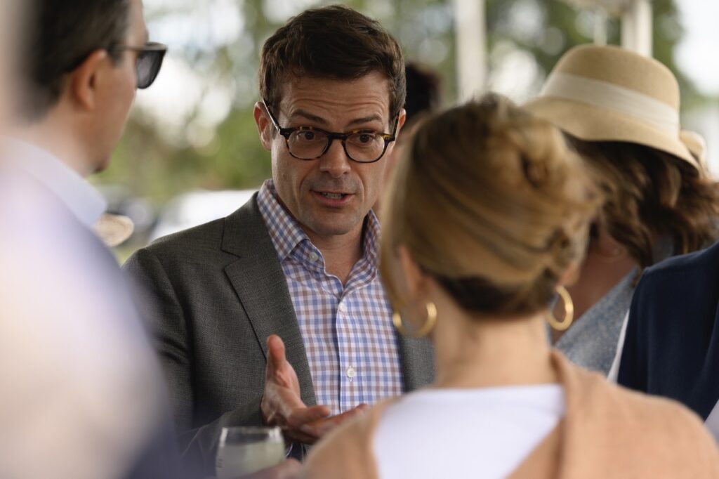 A man in a grey sports coat and brown glasses talks to a woman with gold hoop earrings and a man in sunglasses.