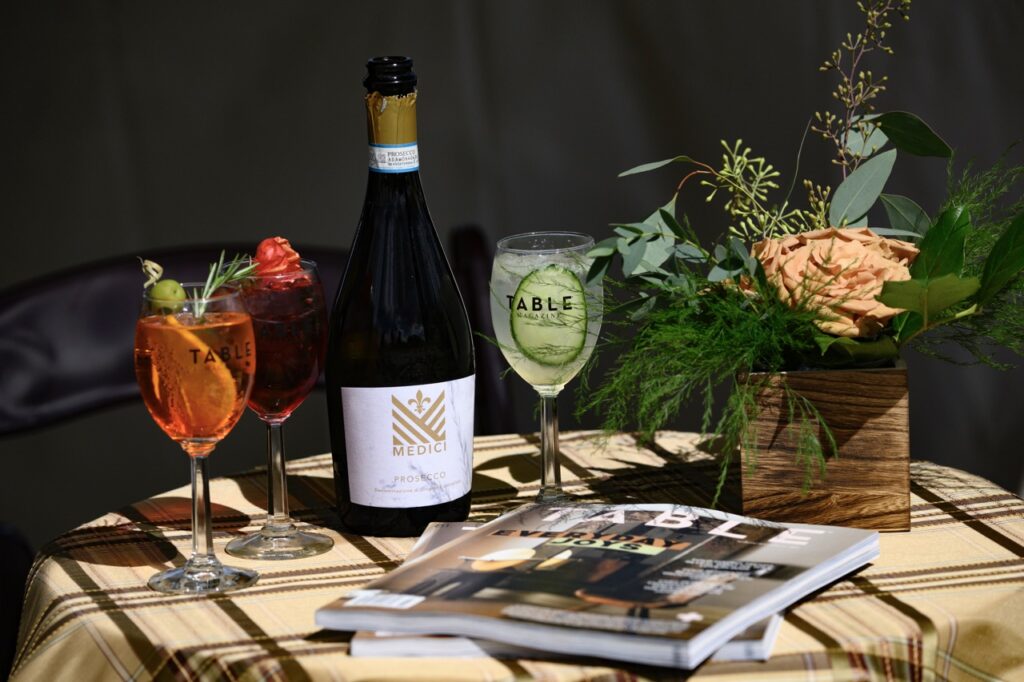 A table set up with TABLE Magazines on the table, a bottle of Medici prosecco, a pink flower arrangement, and three TABLE wine glasses.