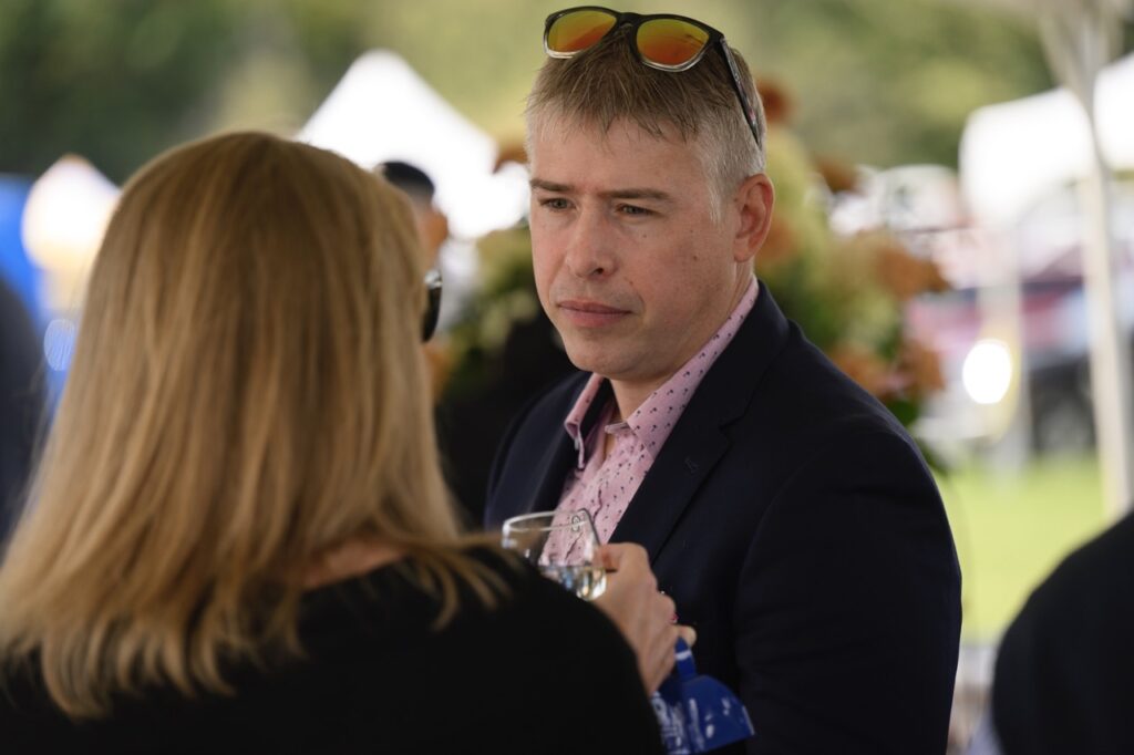A man with sunglasses on his head and a black sports coat talks to a blonde woman with a wine glass in hand.