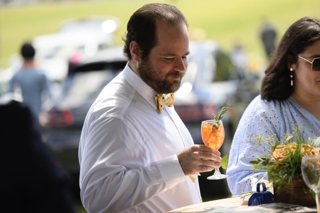 A man with a yellow bowtie and white shirt holds an orange cocktail in a stem glass.