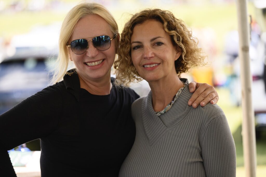 A woman in sunglasses and a black sweater smiles with a woman in a grey sweater with curly blonde hair.