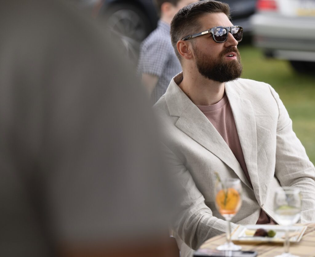 A man in a white sports coat and sunglasses sits at a table with wine glasses.