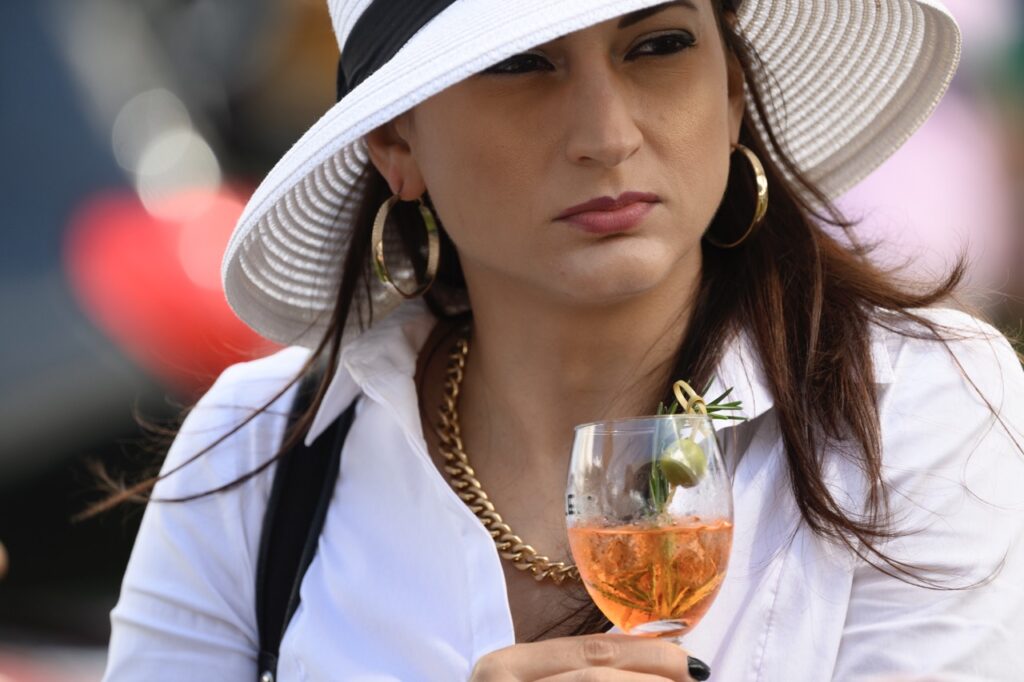 A woman in a white sun hat holds a stem glass with an orange drink and olive inside.
