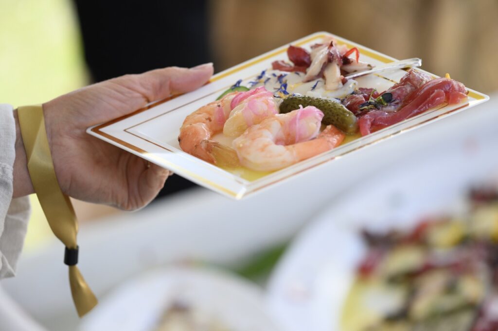 A person holds a small rectangular plate with raw fish, shrimp, small pickles, and veggies.