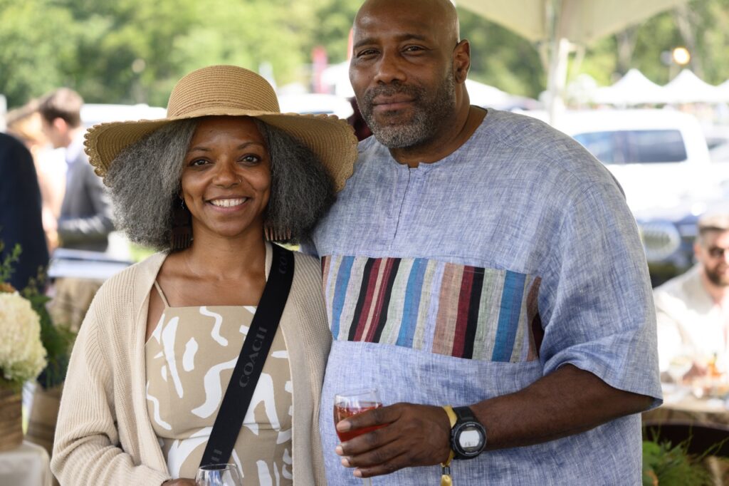 A woman in a brown hat with grey curly hair stands with a man in a blue shirt and wine glass in his hand.
