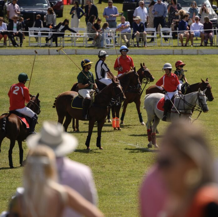 A group of polo players on horses stand on a grassy field with polo sticks on their shoulders.