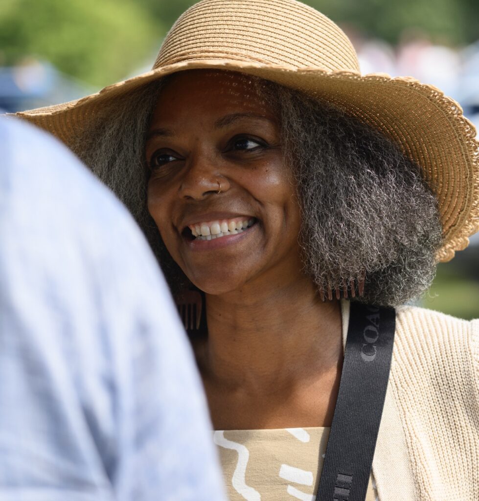 A woman in a brown sun hat with grey hair and a gold nose ring smiles at a man in a blue shirt.