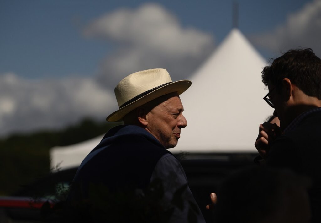A man in a brown hat with a black strip around it stands in the sun in front of a white tent.
