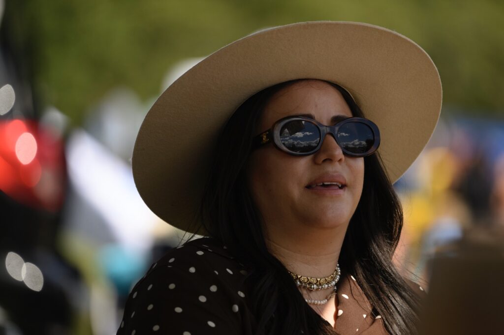 A woman in a brown brim hat and sunglasses looks up at the sky as the clouds reflect in her glasses.