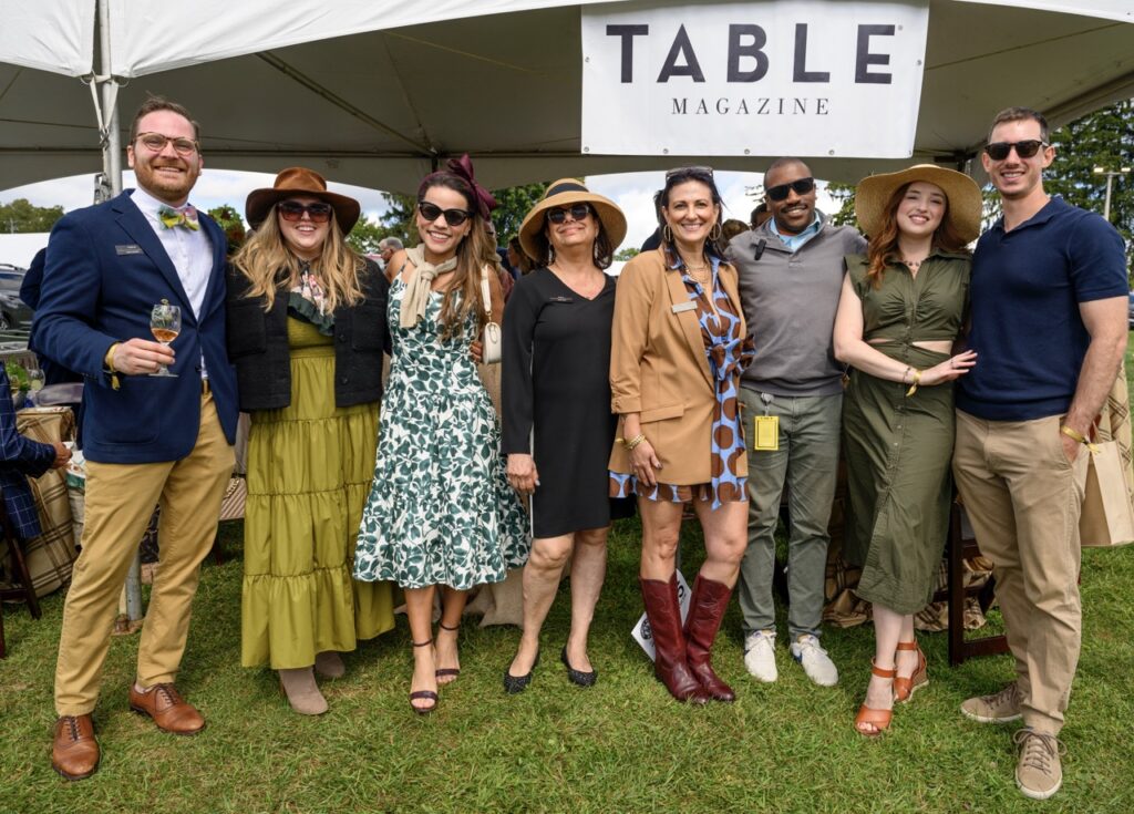A group of people dressed in dresses and hats and button ups and sunglasses stand in a group in front of a TABLE Magazine sign.