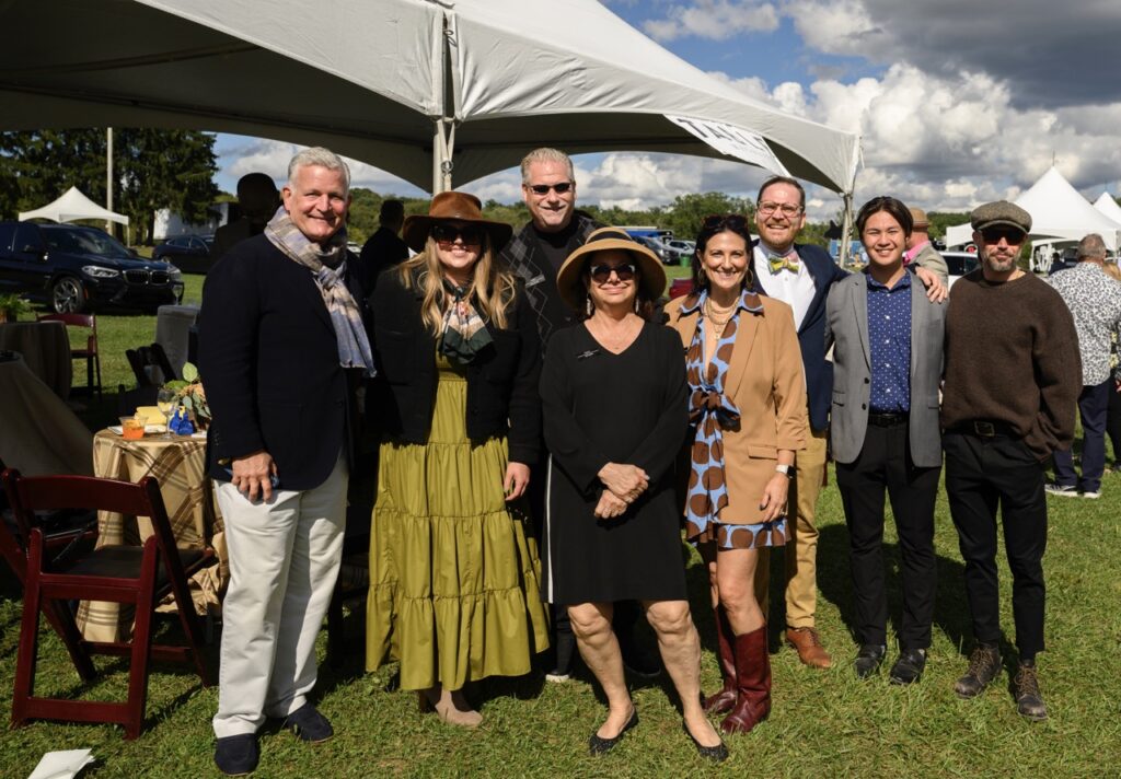 A group of people in dresses, button up shirts, and sweaters stand in a grassy field near a white tent.