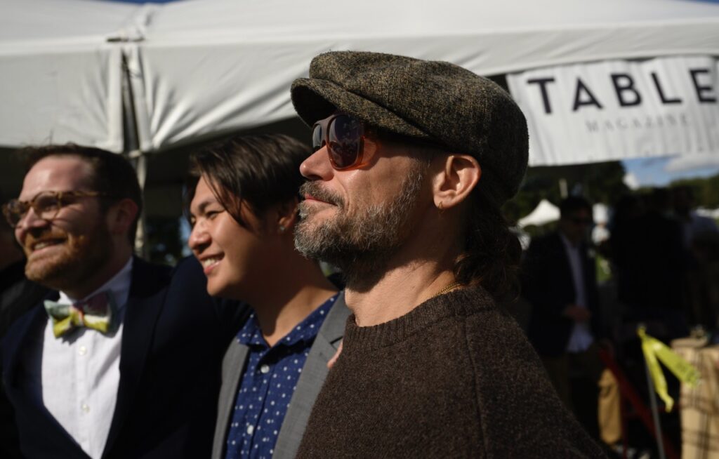A man in a derby hat and sunglasses looks up as the sun as he stands beside two other men.