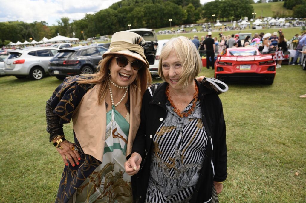 Two women, one in a beige hat smile arm in arm on a grass field surrounded by cars.