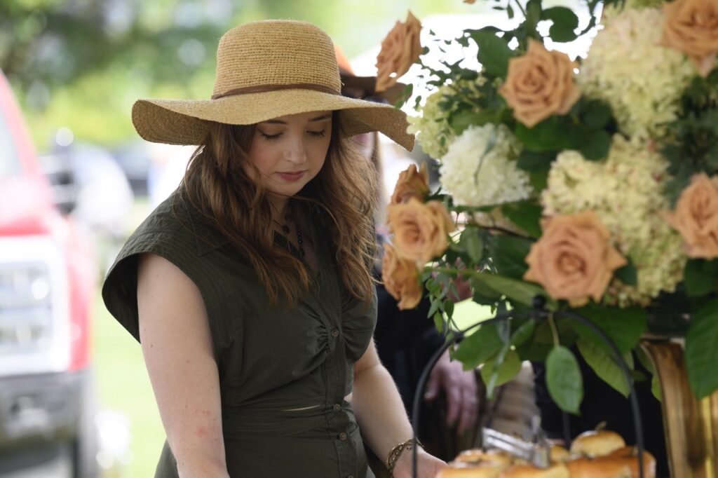 A woman in a brown sun hat and green dress looks down at a table with a huge pink flower arrangement.