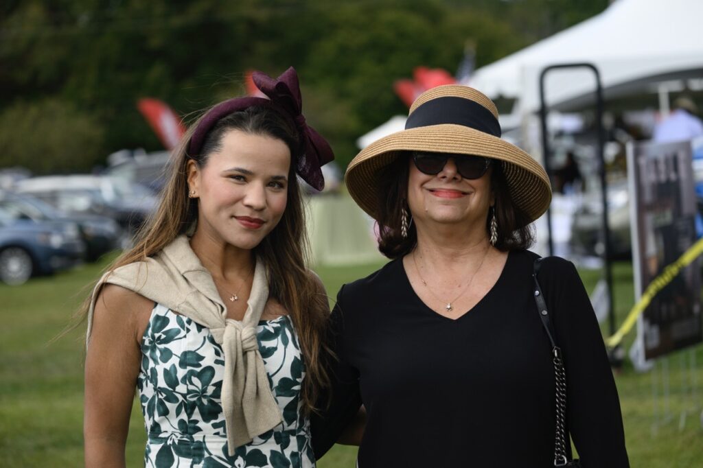 A woman with a purple bow on her head and sweater tied around her shoulders stands with a woman in a brown sun hat, sunglasses, and a black sweater.