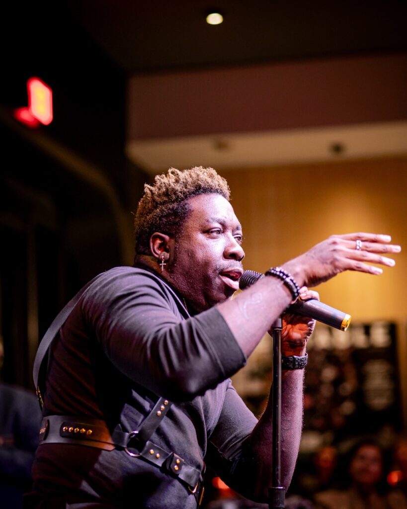 Frzy Winter Party 25 TABLE Magazine Frzy sings on stage into a mic with his arm reaching out at TABLE Magazine's winter preview party.