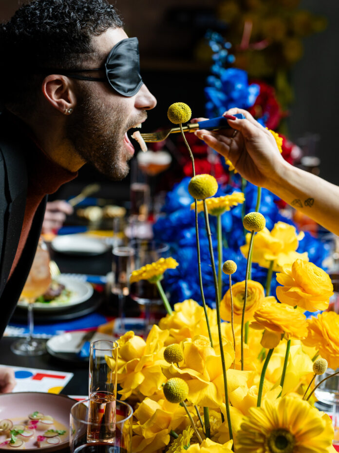A colorful, artistic scene of love is blind, featuring a person wearing a blindfold and surrounded by vibrant yellow flowers, showcasing a creative and sensual approach.