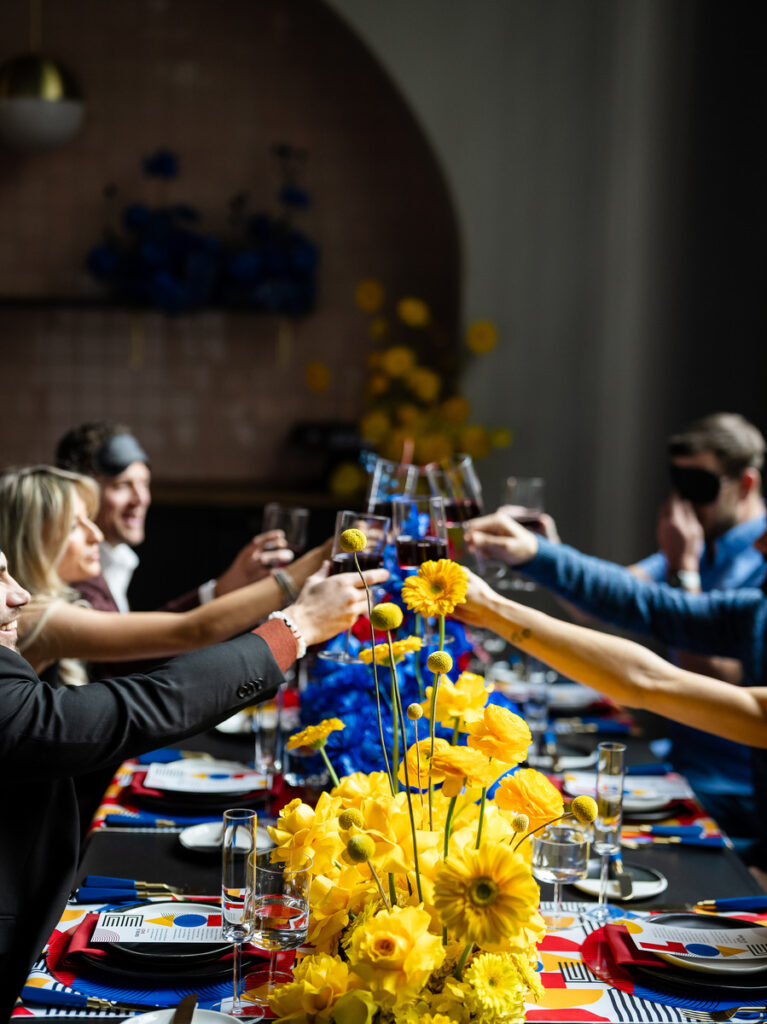 A group of people enjoying a meal at a table decorated with vibrant yellow flowers and colorful geometric patterns, evoking the 'Love is Blind' theme of the event by having some participants wearing blindfolds.