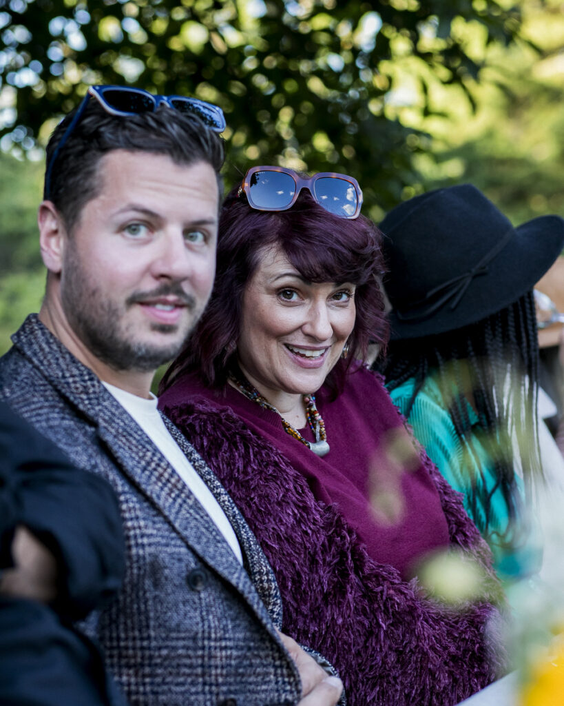 Two individuals, a man and a woman, dressed in festive, stylish attire and posing together outdoors in a lush, green setting.