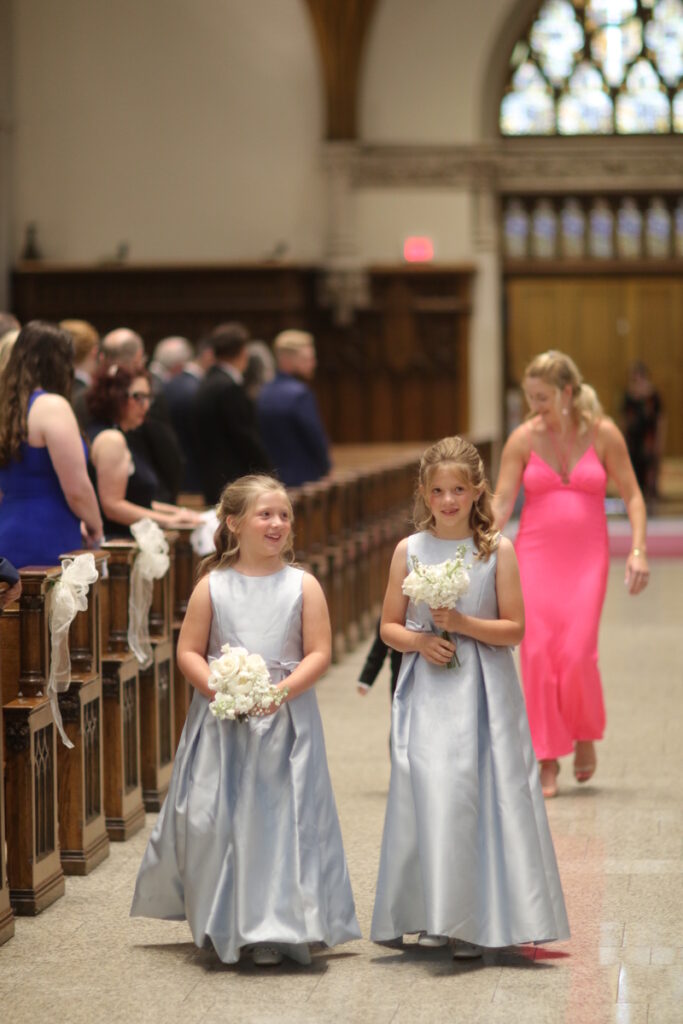 Two young flower girls, dressed in matching silver satin dresses, walking down the aisle of a grand cathedral.