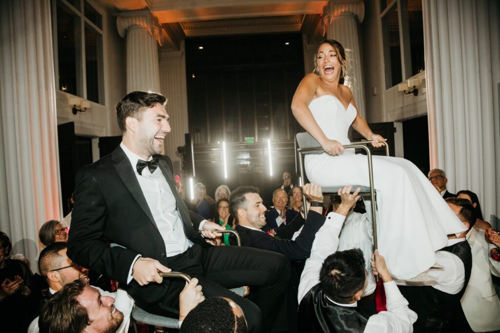 A bride in a white gown gets lifted up in a chair while a groom in a tux gets lifted in a chair as well.